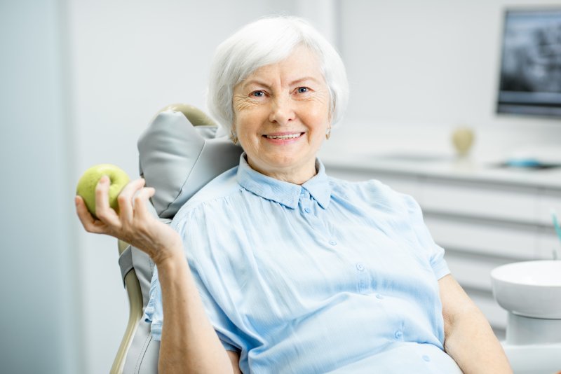 an older woman seated in the dentist’s chair and holding an apple while smiling